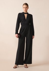 Combinaison noire avec un décolleté en V profond, des manches longues et une taille ceinturée. Tissu lisse, design à jambes larges et détail en forme de trou de serrure au niveau du cou.