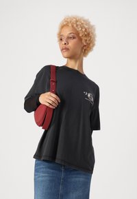 T-shirt noir oversize à manches courtes avec un design graphique, associé à un petit sac bandoulière en cuir rouge, tenu par une sangle.