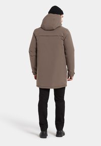 Brun isolerad parka med huva, med en slät yta och längre snitt, kombinerad med svarta byxor och stövlar.
