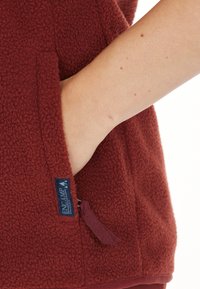 Burgundy fleecemellanmössa med en dragkedjeficka, mjuk textur, rundade kanter och en etikett nära fickan, vilket visar på funktionell design.