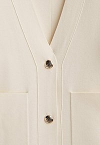 Krämfärgad cardigan med V-ringning, försedd med två framfickor, ribbad textur och en guldfärgad knapp med dekorativ detalj.