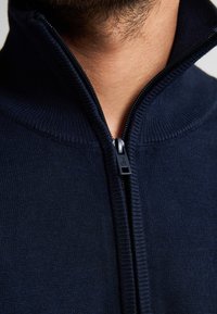 Navy zip-up trui met hoge kraag. Kenmerkt zich door een gebreide textuur en zwarte ritsdetails. Close-up van het halsgebied, geen patronen zichtbaar.