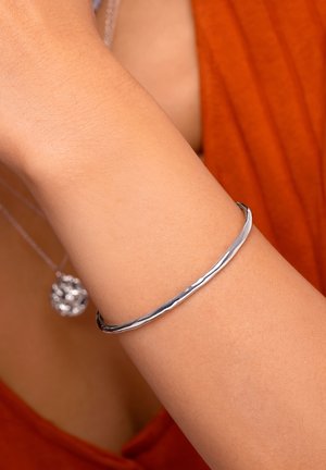 Pulsera de metal plateado con un diseño liso y ligeramente retorcido, llevada en una muñeca sobre un fondo de tela texturizada de color óxido.