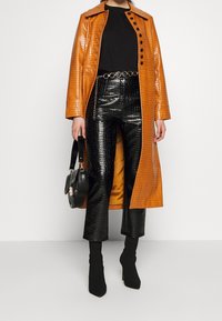 Donna che indossa un lungo cappotto arancione lucido con trama a croc, pantaloni neri con trama a croc, top nero, cintura a catena, borsa nera e stivali neri con tacco.