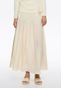 Gonna maxi plissettata di colore avorio, realizzata in un tessuto leggero e testurizzato con cerniera laterale e una leggera lucentezza. Abbinata a sandali beige con zeppa.