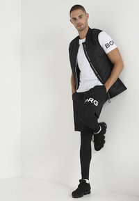 Chaleco acolchado negro con detalle de logo, camiseta blanca, pantalones cortos negros y mallas ajustadas, combinados con zapatillas deportivas negras. Diseño simple, colores monocromáticos.