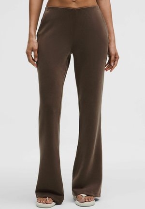 Kobieta ubrana w brązowe spodnie z wysokim stanem i szerokimi nogawkami oraz białe klapki, stojąca na jasnym, jednolitym tle.