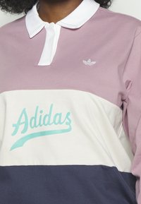 Person in einem farbblockierten Poloshirt in Mauve, Weiß und Marineblau mit einem tealfarbenen "Adidas"-Logo und einem kleinen weißen Adidas-Wappen.