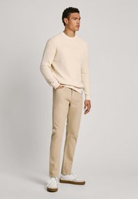 Suéter acanalado de color beige claro, pantalones beige y zapatillas blancas con suelas marrones. El modelo está de pie con las manos en los bolsillos frente a un fondo liso.