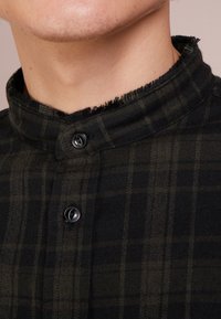 Černá a zelená kostkovaná košile s mandarínovým límcem, roztrhanými okraji a dvěma černými knoflíky na krku. Viditelný detail texturované tkaniny.