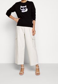 Persona vestida con sudadera negra con un gráfico de gato con gafas de sol, pantalones anchos blancos de cargo y sandalias de tacón abierto blancas, de pie contra una pared simple.