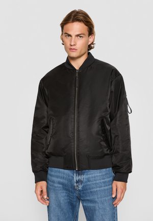 Chaqueta bomber negra hecha de un tejido suave y brillante, con puños y cinturilla acanalados, que presenta bolsillos laterales y cierre con cremallera.
