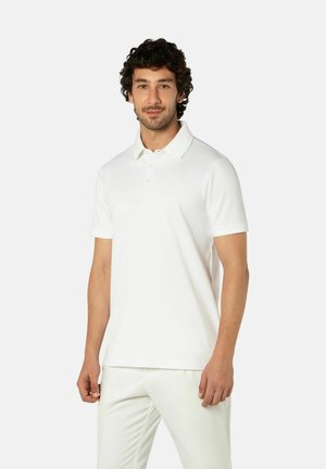 Man die een geheel wit poloshirt met korte mouwen en een broek draagt, staand tegen een effen lichte achtergrond, licht naar rechts gekeerd.