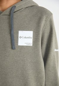 Felpa verde oliva in tessuto texturizzato, con lacci grigi e una toppa col logo Columbia bianco con la scritta "SPORTSWEAR COMPANY" sottostante.