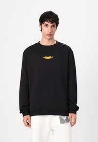 Sort sweatshirt lavet af blødt stof, med et lille gult broderet logo på brystet og ribbede manchetter. Enkel, afslappet design.