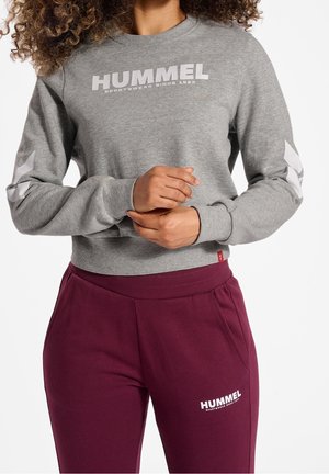 Femme portant un sweat-shirt gris hummel et un pantalon de survêtement hummel bordeaux, mains jointes devant, torse et cuisses visibles.