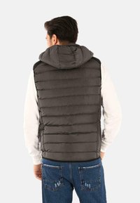 Gilet grigio imbottito con cappuccio, caratterizzato da una trapuntatura orizzontale e un orlo rifinito. Indossato sopra una camicia bianca a maniche lunghe e jeans blu.