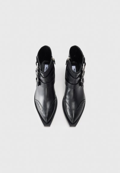 Melnu ādas potītes zābaku pāris ar norādītām pirkstām, dekoratīvām perforācijām, sudraba sprādzēm un sānu rāvējslēdzējiem uz baltas fona.