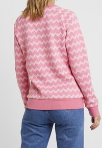 Felpa rosa con motivo a zigzag bianco. Realizzata in tessuto morbido con orlo e polsini a costine, abbinata a jeans blu.