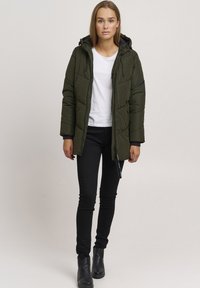 Olive green puffer jacket with a zip front, two side pockets, and a hood. Worn over a white t-shirt and black skinny jeans. Black ankle boots.
