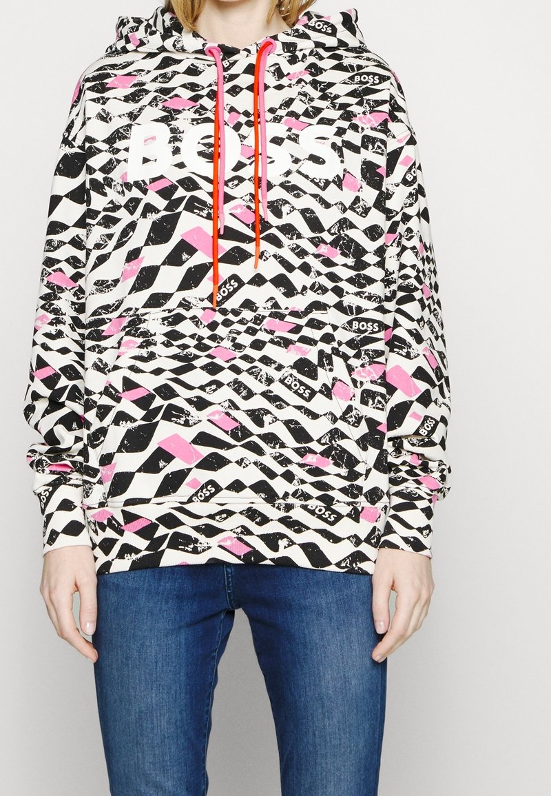 Personne portant un sweat à capuche multicolore noir, blanc et rose avec des cordons rouges et le texte « BOSS », associé à un jean bleu.