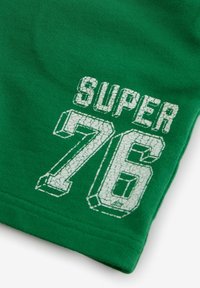 Grön tyg med en texturerad vit tryck där texten "SUPER 76" i blockstil är placerad på den nedre delen av plagget.
