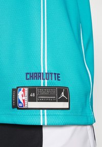 Sinih koripallo-asu, jossa on valkoiset raidat ja jossa lukee "CHARLOTTE" paksuilla purppurakirjaimilla. Mukana musta etiketti, jossa on NBA:n brändäys ja koko 48.