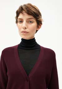 Bordeaux kleurige cardigan met een V-hals, ribbeltextuur en een enkele knoopsluiting, opgedragen over een zwarte coltrui.