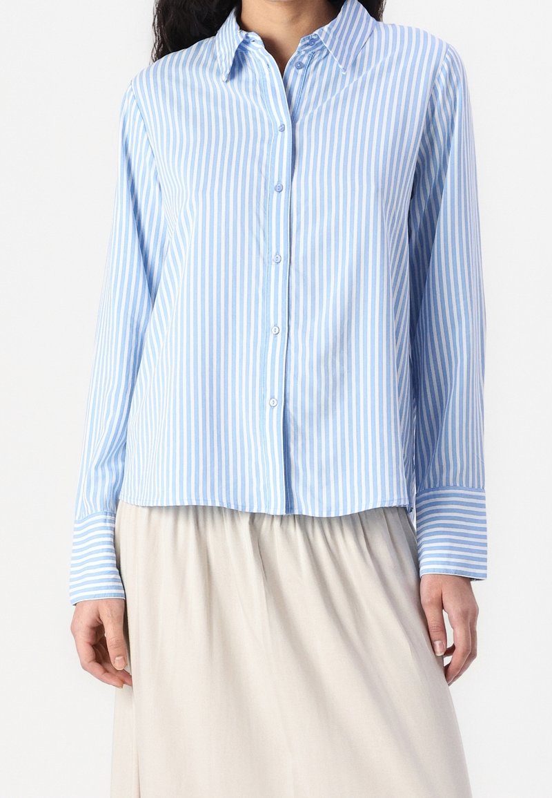 Femme portant une chemise à rayures verticales bleu clair et blanches à manches longues et une jupe beige, debout devant un fond uni.