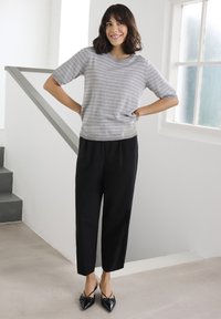 Hall triibuline T-särk lühikeste varrukatega, mustade lühendatud pükstega ja teravate mustade kingadega, seistes heledas interjööris.