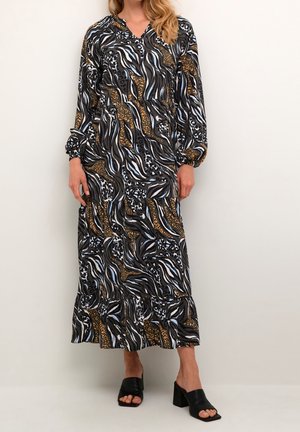 Femme portant une robe longue à manches longues, longueur cheville, avec un imprimé animal abstrait noir, blanc, bleu et marron, et des chaussures ouvertes à talons noirs.