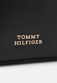 Czarna syntetyczna torebka z metalowym złotym logo z napisem "TOMMY HILFIGER" na fakturowanej powierzchni. Prosty, elegancki design z wyraźnym logo.