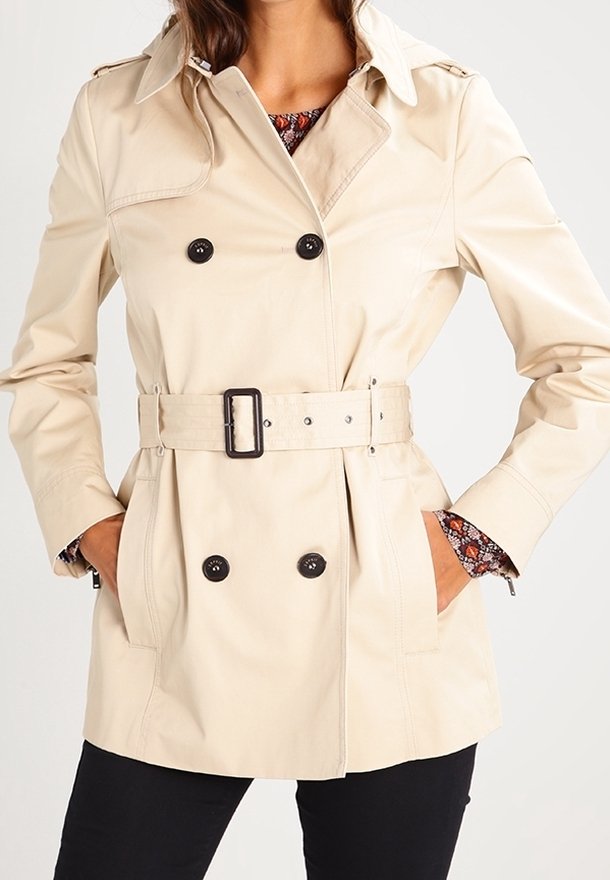 Trench-coat beige croisé avec boutons noirs et ceinture, porté sur une chemise intérieure fleurie, associé à un pantalon noir, mains dans les poches.