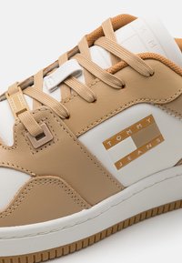 Baskets beige et blanches avec lacets beiges et logo Tommy Jeans sur le côté, avec des panneaux cousus et une semelle en caoutchouc texturée.