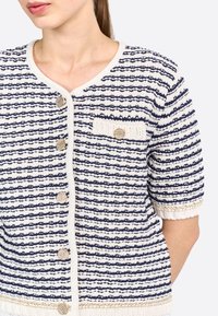 Gilet en tricot avec un motif rayé bleu marine et crème, à manches courtes, col rond, boutons dorés, et une petite poche avec une patch.