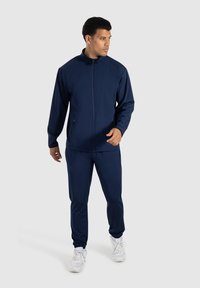 Námořnický teplákový komplet z hladkého materiálu, obsahující zipovou bundu s dvěma bočními kapsami a odpovídající zúžené kalhoty, doplněné bílými teniskami.