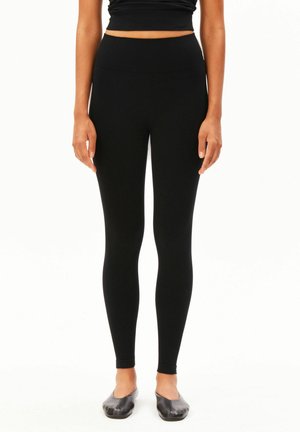 Vrouw draagt zwarte high-waist leggings, zwart crop top en zwarte ballerina's, staande tegen een witte achtergrond.