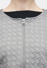 Κοντινό πλάνο γκρι μπουφάν με ραφές και ασημένιο φερμουάρ, μερικώς κλειστό στο λαιμό πάνω από μαύρο εσωτερικό πουκάμισο.