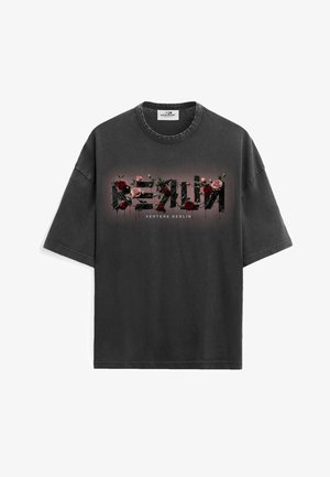 Jeune homme aux cheveux courts portant un t-shirt noir délavé oversize avec un motif floral et un pantalon blanc, debout les mains dans les poches.