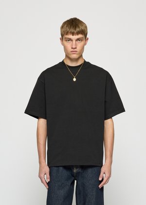 Young man with short brown hair wearing a loose black t-shirt, blue jeans, and a gold pendant necklace, standing against a plain background.