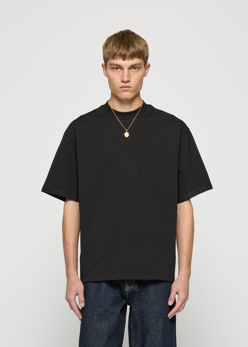 Young man with short brown hair wearing a loose black t-shirt, blue jeans, and a gold pendant necklace, standing against a plain background.