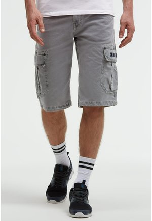 Mann trägt graue Cargoshorts mit Seitentaschen, weiße Socken mit schwarzen Streifen und schwarze Turnschuhe, steht vor neutralem Hintergrund.