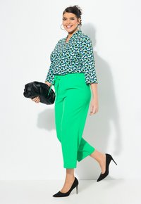 Zelené kalhoty na míru s páskem a vzorovaná blůza v modré, zelené a bílé barvě, doplněné černými botami na podpatku s špicí. Drží černou klutch.