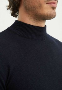 Pull à col montant côtelé bleu marine foncé, fabriqué à partir d'un matériau doux, avec un col haut et une texture lisse. Gros plan sur les détails du tissu.