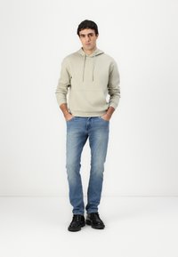 Sweatshirt cinza claro com bolso canguru e cordão, combinado com jeans azuis desbotados e sapatos pretos, de pé sobre um fundo simples.