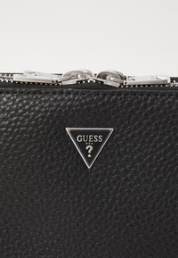 Billetera negra textura con un cierre plateado, que presenta un acento del logo triangular de GUESS. El material parece de cuero, con una superficie estampada.