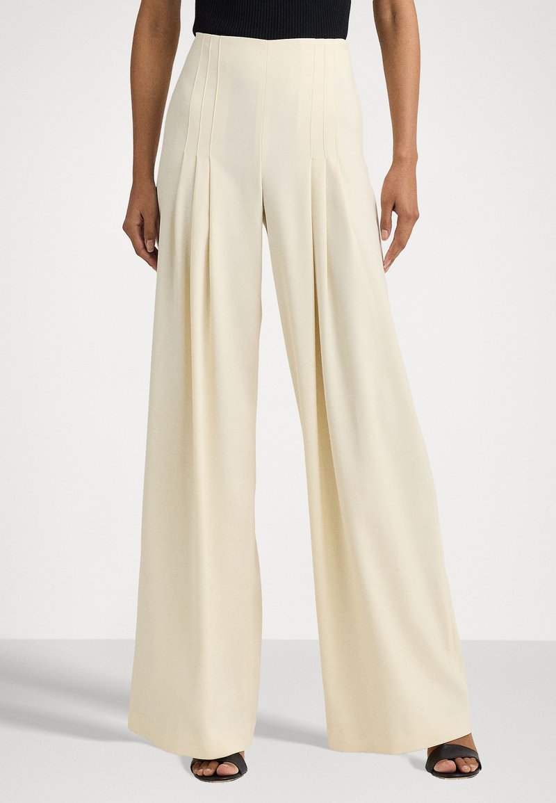 Nainen, jolla on ylhäisiin vyötäröihin ulottuvat leveälahkeiset kermaiset housut etumaisilla poimutuksilla ja mustat avonaiset kengät, seisoo yksinkertaista taustaa vasten.