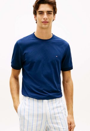 Giovane uomo che indossa una t-shirt blu navy tinta unita e pantaloni bianchi con sottili righe verticali blu, in piedi con le mani in tasca.