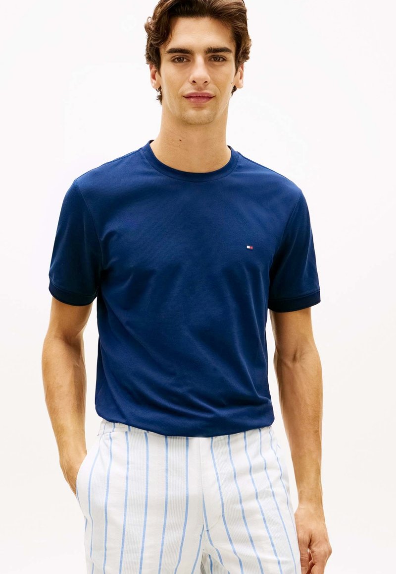 Jeune homme portant un t-shirt bleu marine uni et un pantalon blanc avec de fines rayures verticales bleues, debout les mains dans les poches.