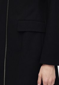 Κοντινή λήψη ενός μαύρου παλτού με μπροστινό φερμουάρ και λεπτομέρεια τσέπης με καπάκι, που δείχνει το χέρι ενός ατόμου να ακουμπά δίπλα στο μανίκι.